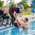 A young man is being helped from a wheelchair into a swimming pool by two trainers.